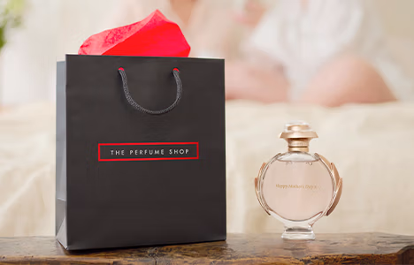 A bottle of Paco Rabanna Olympea on a wooden ledge, next to a black gift bag with The Perfume Shop logo on it