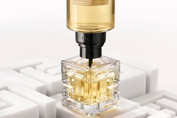 Balmain perfume bottle with gold liquid dispensing into a sculpted glass bottle, styled on a white geometric background