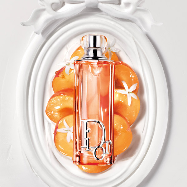 Dior Addict peach-toned perfume bottle displayed on a white ornate frame with sliced peach accents and small white flowers.