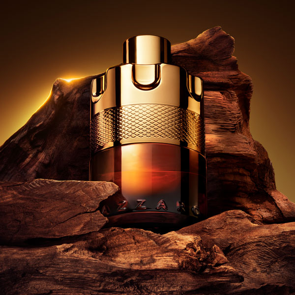 Azzaro Forever Wanted Absolu fragrance bottle with a polished gold cap and textured gold band, fading into amber glass at the base. The bottle is nestled between rugged dark rocks under warm golden lighting, with the engraved Azzaro name visible near the bottom.