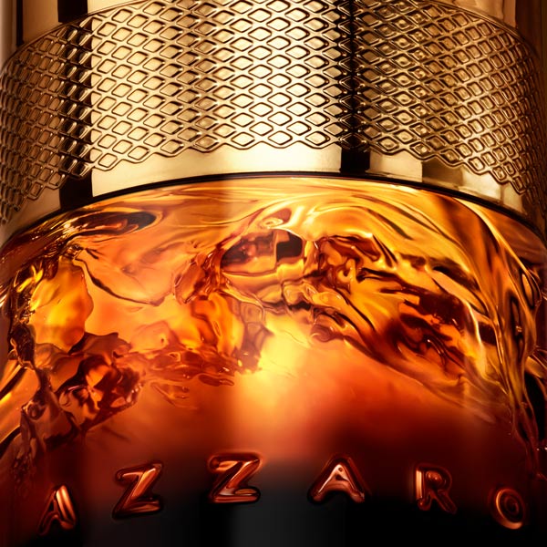 Extreme close-up of the lower half of an Azzaro Forever Wanted Absolu fragrance bottle, showing swirling amber liquid behind thick glass beneath a textured gold metal band. Warm golden lighting highlights the liquid’s whisky-like tones, with the embossed AZZARO lettering visible at the base.