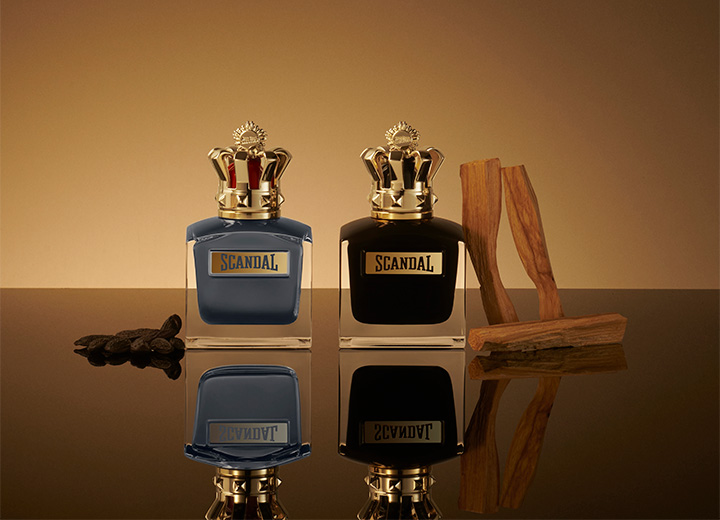 Two Scandal perfume bottles in blue and black with gold crown caps, styled with tonka beans and wood on a reflective surface.