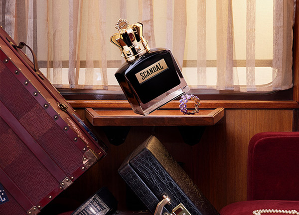 Scandal perfume bottle with gold crown cap displayed on a wooden shelf among vintage suitcases and accessories.