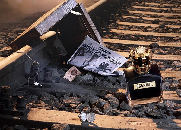 Scandal perfume bottle on railway tracks beside an open suitcase and vintage newspaper in a dramatic, smoky setting.