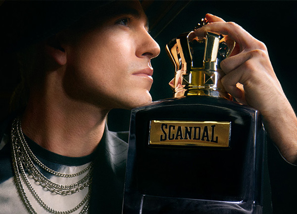Close‑up of a Scandal perfume bottle with a gold crown cap, held beside layered chain jewellery in a dark, dramatic setting.