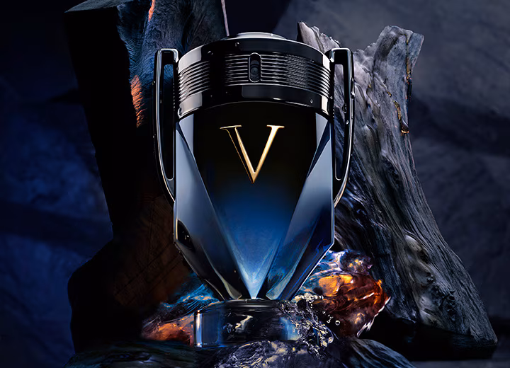 V‑shaped perfume bottle with a gold V emblem, set on dark rocks with blue and amber tones, highlighting an intense, masculine fragrance.